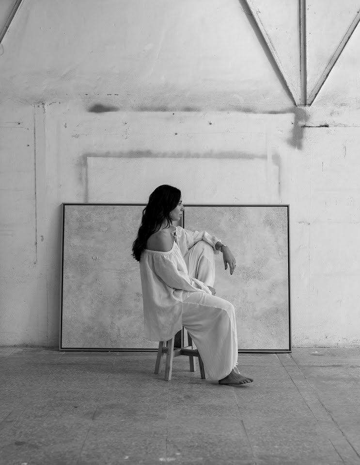 Irene sentada de perfil en un taburete, delante de dos cuadros con marco de madera, en blanco y negro